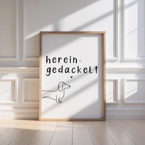 May include: A framed print featuring the words "herein-gedackelt" in black script above a minimalist line drawing of a dachshund. The artwork is set against a white background and framed in light brown wood.