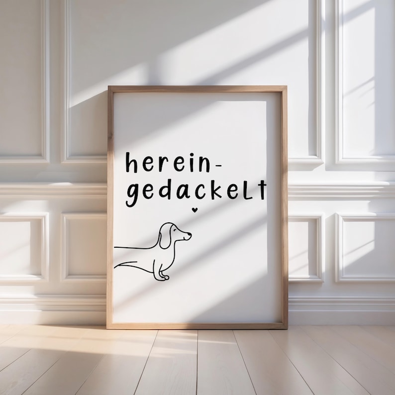 May include: A framed print featuring the words "herein-gedackelt" in black script above a minimalist line drawing of a dachshund. The artwork is set against a white background and framed in light brown wood.