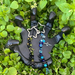 May include: Two star-shaped keychains and two beaded keychains with the letters "JUCK" are displayed on a black hand-shaped stand. The beaded keychains feature green, black, and blue beads. One keychain has a star charm.