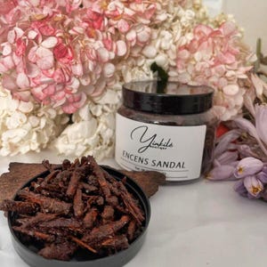 Può includere: Un barattolo di vetro con un coperchio nero pieno di trucioli di incenso marroni. Il barattolo ha un'etichetta bianca con il testo "Yinkile Boutique" e "Encens Sandal".