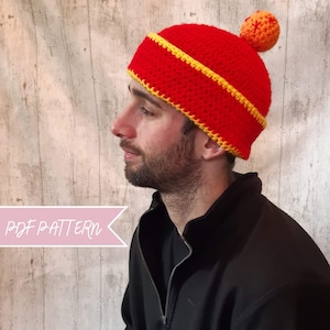 Puede incluir: Un gorro de punto rojo y amarillo con una pequeña bola naranja de punto en la parte superior. El gorro tiene una franja amarilla alrededor del borde. El gorro lo lleva puesto una persona con barba.
