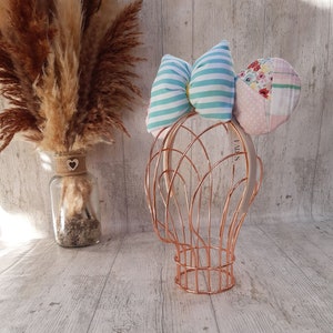 Puede incluir: Una diadema con dos lazos grandes. Un lazo es azul y blanco a rayas y el otro es rosa y con estampado floral. La diadema está en un soporte de alambre de cobre.