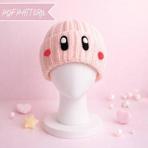 Puede incluir: Un gorro de punto rosa claro con un diseño de cara de dibujos animados, con ojos negros y mejillas rojas. El texto "PDF PATTERN" está en la esquina superior izquierda. El sombrero se muestra en una cabeza de maniquí blanca.