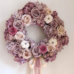 May include: A circular wreath featuring an arrangement of mauve, dusty rose, and cream-colored artificial roses and hydrangeas interspersed with small white and dark pink pumpkins. A light beige ribbon hangs from the bottom center of the wreath.