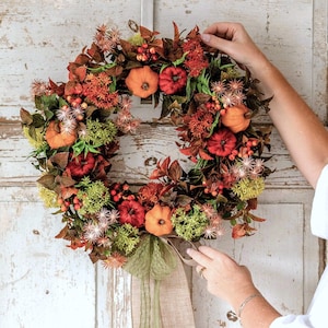 May include: A decorative autumn wreath featuring orange and red pumpkins, brown leaves, green foliage, and small red berries. The circular arrangement is accented with spiky, star-shaped floral elements and a light green sheer ribbon tied at the bottom.