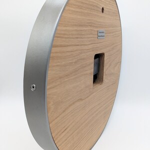 May include: A round wooden clock face with a silver metal frame. The clock face has a square opening for the clock mechanism. The back of the clock has a small metal plate with the text "Handcrafted by Modern Furniture".