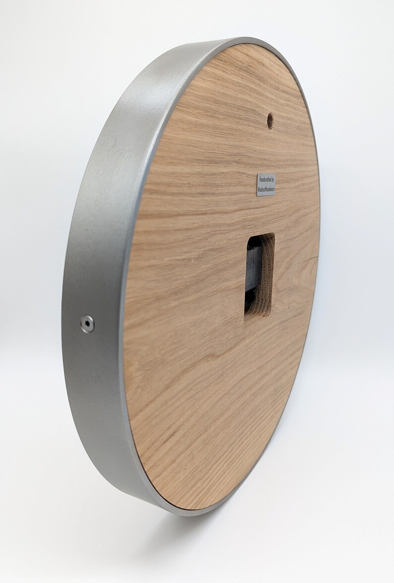 May include: A round wooden clock face with a silver metal frame. The clock face has a square opening for the clock mechanism. The back of the clock has a small metal plate with the text "Handcrafted by Modern Furniture".