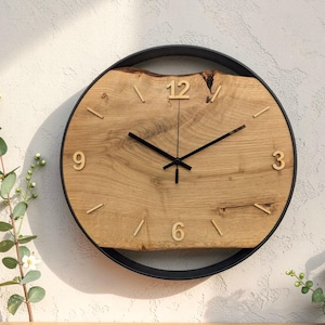 Könnte beinhalten: Eine runde Wanduhr mit einem Zifferblatt aus einer hellbraunen Holzscheibe mit Naturkante, eingefasst in einen dünnen, schwarzen, kreisförmigen Metallrahmen. Das Zifferblatt zeigt goldene Ziffern bei 12, 3, 6 und 9 sowie schwarze Zeiger.