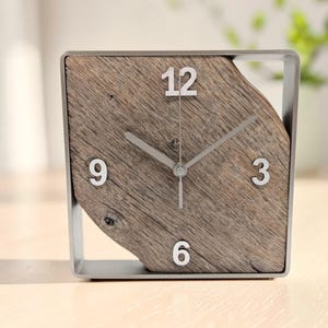 Könnte beinhalten: Eine quadratische Uhr mit einem Zifferblatt aus natürlichem, verwittertem braunem Holz mit sichtbarer Maserung. Die Uhr ist in einen dünnen, silberfarbenen Metallrahmen gefasst. Sie zeigt silberne Ziffern an den Positionen 12, 3, 6 und 9 sowie passende Zeiger.