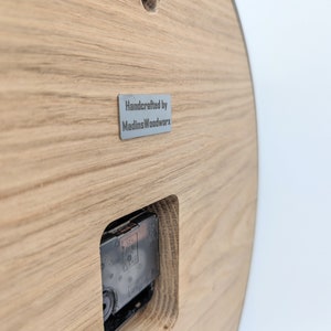 May include: The back of a wooden clock with a silver metal label that reads "Handcrafted by MadinsWoodworx".