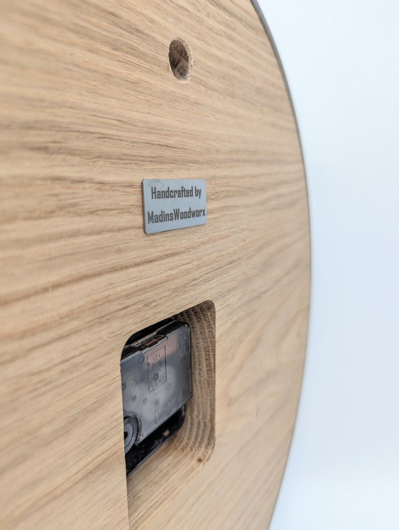 May include: The back of a wooden clock with a silver metal label that reads "Handcrafted by MadinsWoodworx".