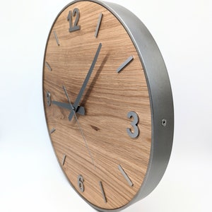 May include: A round wooden clock with a silver metal frame and silver hands. The clock face has Roman numerals for the hours and silver lines for the minutes. The clock is 12 inches in diameter.