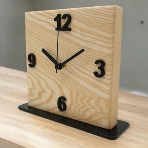 Könnte beinhalten: Eine Holzblockuhr mit schwarzen Ziffern und Zeigern. Die Uhr hat eine helle Holzmaserung und steht auf einem schwarzen rechteckigen Sockel. Die Zahlen 3, 6, 9 und 12 sind sichtbar.