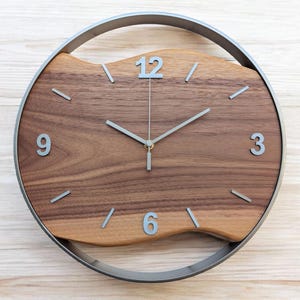 Könnte beinhalten: Eine runde Wanduhr mit Holzgesicht und silbernem Metallrahmen. Das Zifferblatt ist aus hellbraunem Holz mit silbernen Ziffern und Zeigern. Der Durchmesser beträgt ca. 30 cm.