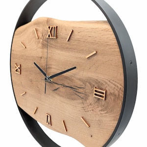 May include: A round, black metal frame surrounds a wooden clock face with Roman numeral markings. The clock face is made of a natural wood slice with a unique grain pattern.