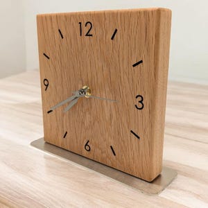 Könnte beinhalten: Eine quadratische Holzuhr mit hellbraunem Finish. Das Zifferblatt hat schwarze Ziffern und Striche, die die Stunden markieren. Silberne Zeiger zeigen die Uhrzeit an. Die Uhr steht auf einem silbernen Metallsockel.