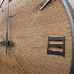 May include: A large, round, wooden clock with Roman numeral markings and black metal hands. The clock face is made of light brown wood with a natural grain pattern. The outer edge of the clock is a silver metal ring.