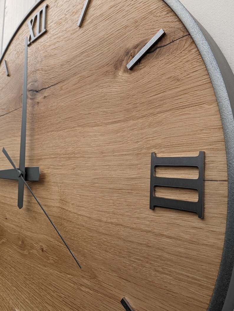 May include: A large, round, wooden clock with Roman numeral markings and black metal hands. The clock face is made of light brown wood with a natural grain pattern. The outer edge of the clock is a silver metal ring.