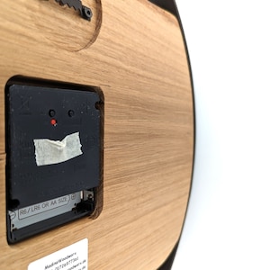 May include: The back of a wooden clock with a black clock mechanism. The clock is powered by AA or LR6 batteries. The clock has a label with the text "Madins Woodworx" and contact information.