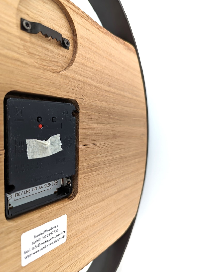 May include: The back of a wooden clock with a black clock mechanism. The clock is powered by AA or LR6 batteries. The clock has a label with the text "Madins Woodworx" and contact information.
