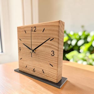 Könnte beinhalten: Eine quadratische Holztischuhr mit hellbrauner Holzmaserung. Das Zifferblatt zeigt schwarze Ziffern an den Positionen 12, 3, 6 und 9, schwarze Striche für die anderen Stunden sowie schwarze Zeiger. Die Uhr steht auf einem rechteckigen schwarzen Sockel.