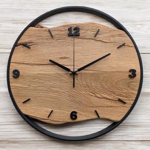 Könnte beinhalten: Runde Wanduhr mit hellem Holz-Zifferblatt und schwarzen Zeigern und Ziffern. Das Zifferblatt aus hellem Holz hat einen unregelmäßigen Rand. Die Uhr ist von einem schwarzen Metallrahmen umgeben. Der Durchmesser beträgt ca. 30 cm.