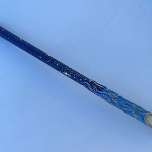 Puede incluir: Una varita azul y dorada con un diseño de cielo estrellado. La varita está hecha de madera y tiene una punta puntiaguda.