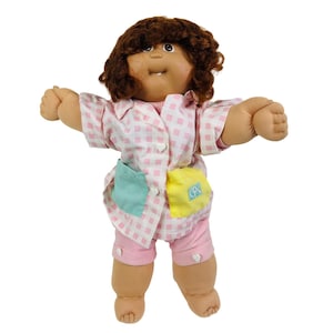 May include: A vintage Cabbage Patch Kid doll with brown curly hair and a smiling expression. The doll wears a pink and white checkered pajama top and shorts with a light blue and yellow pocket. The doll's arms are outstretched.