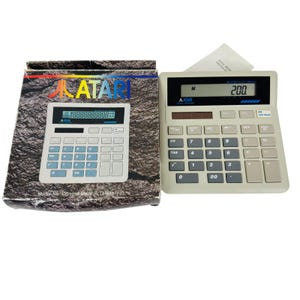 May include: An Atari CC1010 calculator with its original box. The calculator is light gray with a black display showing "200". The box has the Atari logo and a picture of the calculator. The text "Model No. CC1010 Made in Taiwan" is visible.