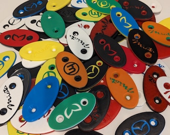 May include: A collection of colorful oval-shaped keychains with various logos and text, including "Mazda", "Miata", and "Sport".