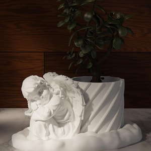 May include: A white ceramic planter with a sculpted angel design, holding a small green plant. The planter has a textured, spiral design and sits on a marble surface. The angel is in a seated position with its head resting on its knees.