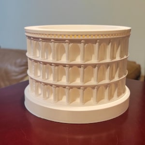 May include: A three-tiered, off-white model of the Colosseum. The miniature replica features arched openings and columns, with a circular base and top. The model is a detailed representation of the historic Roman amphitheater.