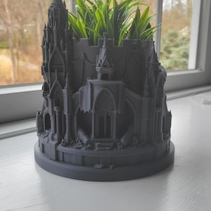 May include: A gray, 3D-printed castle-shaped planter with a round base. The planter features intricate architectural details, including turrets, arches, and decorative elements. Green plants are visible inside the planter, adding a touch of nature to the design.