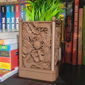 May include: A brown rectangular planter with intricate dragon carvings. The planter holds vibrant green artificial plants. The design features detailed relief work, creating a textured appearance. The planter is placed on a shelf with books in the background.