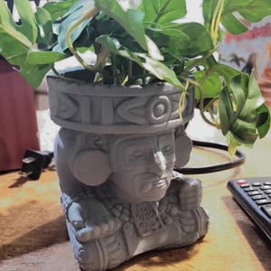 May include: A gray Aztec-style planter pot with a detailed face and headdress design, filled with green leafy plants. The planter is made of a solid material and sits on a table. The plants add a natural element to the decorative pot.