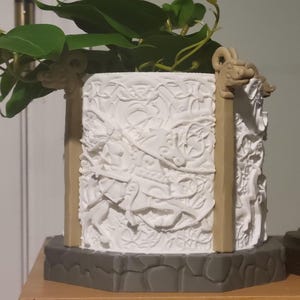 May include: A white, square planter with intricate relief carvings and tan-colored supports. The planter sits on a gray base and holds green leafy plants. The design features dragon-like figures.