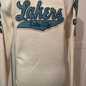 May include: White knitted sweater with a blue felt patch that says "Lakers" and "Lake City" on the back.