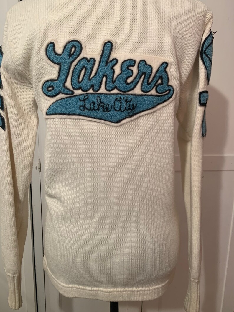 May include: White knitted sweater with a blue felt patch that says "Lakers" and "Lake City" on the back.
