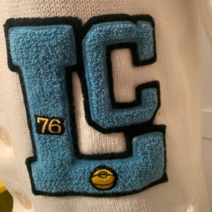May include: A white knitted cardigan with a blue and black felt patch that says "LC" with the number "76" in a smaller font. A yellow felt basketball is on the patch.