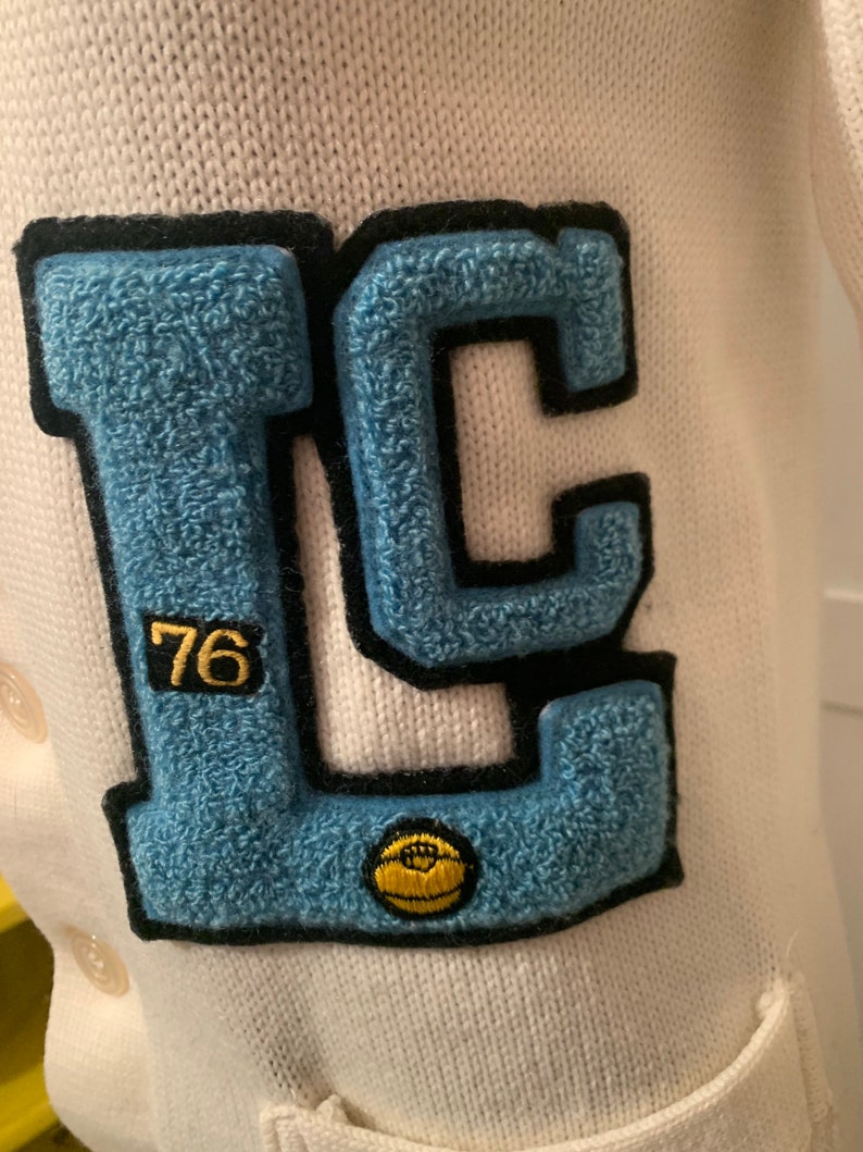 May include: A white knitted cardigan with a blue and black felt patch that says "LC" with the number "76" in a smaller font. A yellow felt basketball is on the patch.