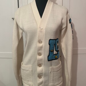 May include: A white cardigan sweater with a blue and white embroidered patch on the left side. The patch has the letters "L" and "C" and the number "76".