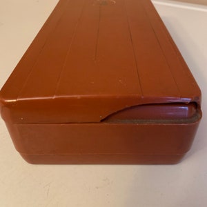 Puede incluir: Una caja rectangular de pl&aacute;stico marr&oacute;n con una esquina ligeramente da&ntilde;ada. La caja tiene una parte superior acanalada y una peque&ntilde;a hendidura circular en la superficie superior. El color es un marr&oacute;n rojizo c&aacute;lido.