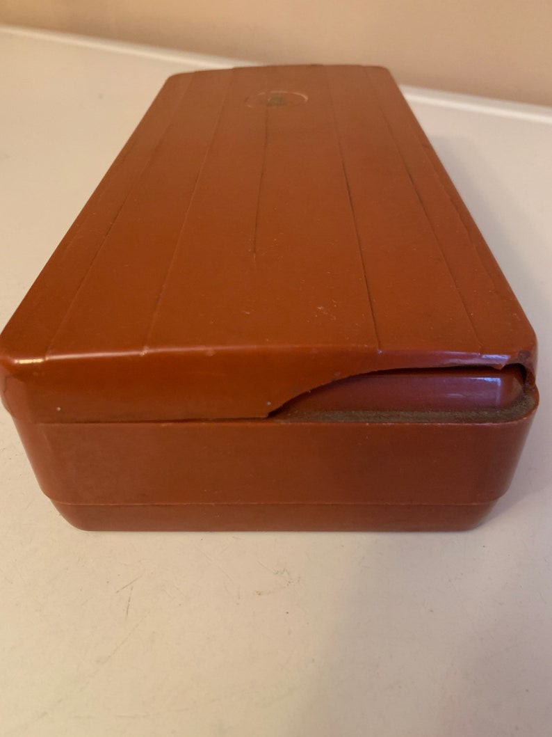 Puede incluir: Una caja rectangular de pl&aacute;stico marr&oacute;n con una esquina ligeramente da&ntilde;ada. La caja tiene una parte superior acanalada y una peque&ntilde;a hendidura circular en la superficie superior. El color es un marr&oacute;n rojizo c&aacute;lido.