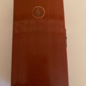 Puede incluir: Un estuche rectangular de color marr&oacute;n rojizo con estr&iacute;as verticales. Un peque&ntilde;o emblema circular con un dise&ntilde;o dorado se encuentra cerca de la parte superior. El estuche tiene un cierre negro en el lado derecho.