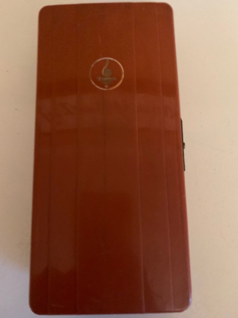 Puede incluir: Un estuche rectangular de color marr&oacute;n rojizo con estr&iacute;as verticales. Un peque&ntilde;o emblema circular con un dise&ntilde;o dorado se encuentra cerca de la parte superior. El estuche tiene un cierre negro en el lado derecho.