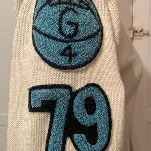 May include: White knitted sweater with a blue and black felt patch on the sleeve. The patch features a circle with the letter "G" and the number "4" inside. Below the patch is a blue and black felt number "79".
