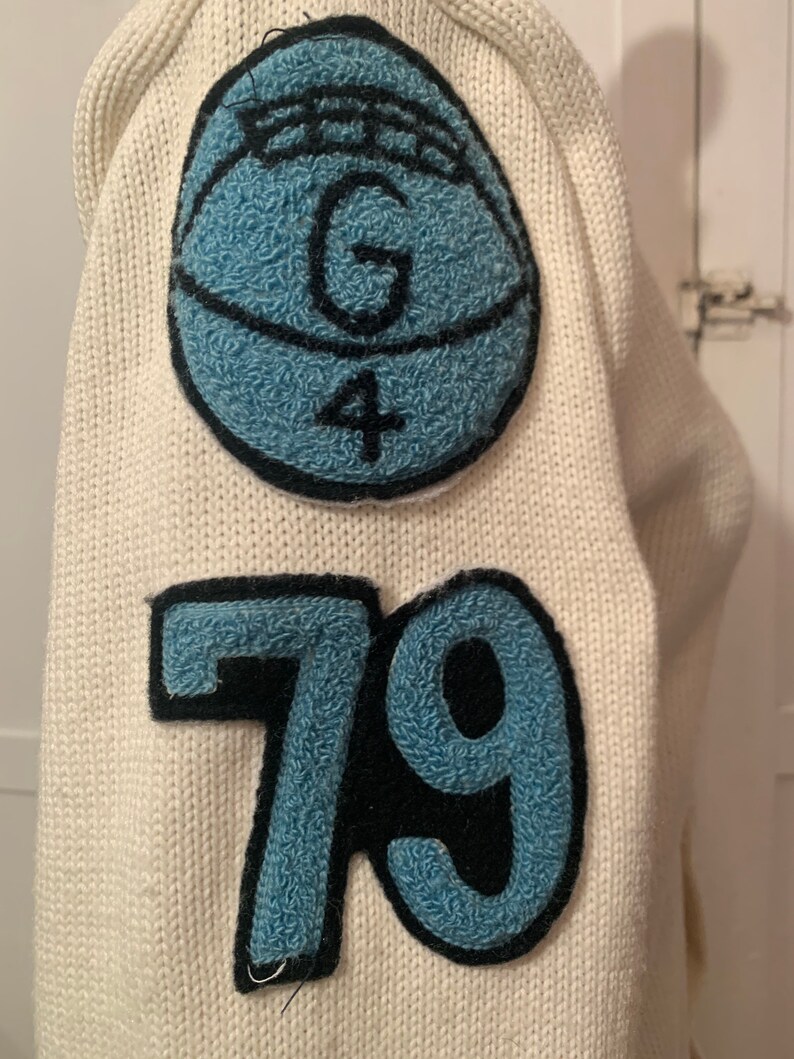 May include: White knitted sweater with a blue and black felt patch on the sleeve. The patch features a circle with the letter "G" and the number "4" inside. Below the patch is a blue and black felt number "79".