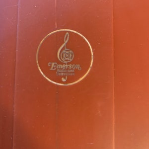 Puede incluir: Primer plano de una superficie marr&oacute;n con un emblema dorado. El emblema presenta una nota musical y la palabra "Emerson" en una fuente estilizada, con texto adicional debajo. El dise&ntilde;o est&aacute; encerrado en un c&iacute;rculo dorado.