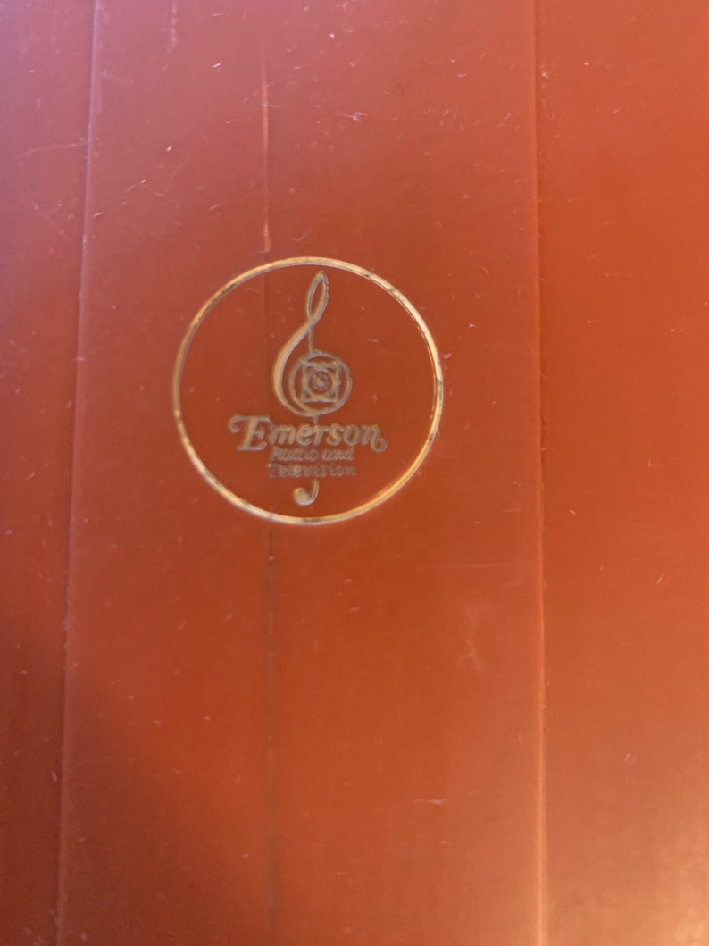 Puede incluir: Primer plano de una superficie marr&oacute;n con un emblema dorado. El emblema presenta una nota musical y la palabra "Emerson" en una fuente estilizada, con texto adicional debajo. El dise&ntilde;o est&aacute; encerrado en un c&iacute;rculo dorado.