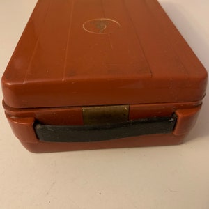 Puede incluir: Un estuche rectangular marr&oacute;n con asa negra y cierre de lat&oacute;n. La caja tiene una textura acanalada en la parte superior y un peque&ntilde;o emblema ovalado. La caja est&aacute; cerrada y parece estar hecha de pl&aacute;stico duro o material compuesto.